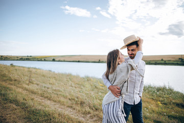 Fototapeta premium Happy couple in love hugging, kissing and smiling against the sky in field. Hat in girl's hand