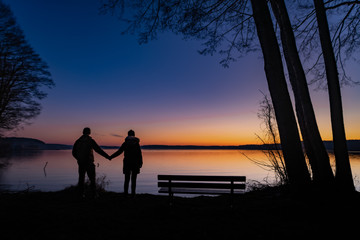 Wieczorny zachód słońca nad jeziorem. Para trzymająca się za ręce i ławka
