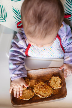 Baby girl sitting in high chair eating homemade oatmeal cookies with hands, Top view