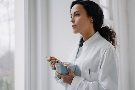 Female doctor eating a snack, standing at window