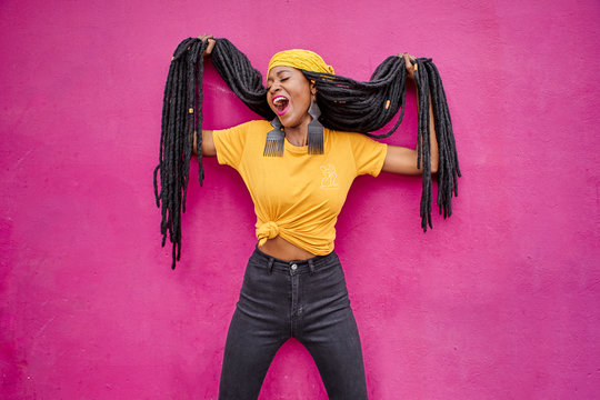 Portrait of woman with long dreadlocks holding her hair in front of a pink wall