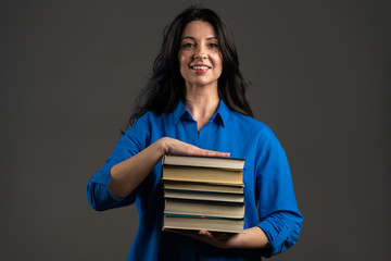 European smiling mature woman as student on grey background in studio holds stack of university books from library. Adult lady smiles, she is happy to graduate.
