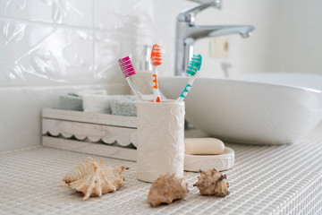 Three different colored toothbrushes stand in a ceramic glass against the background of the washbasin and other accessories in the bathroom