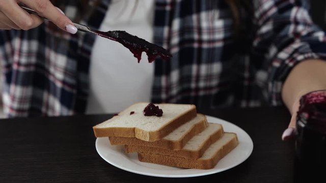 Young woman in kitchen duringquarantine. Close up motion of woamn using fork for putting some red jam on bread. Sweet toast ready to be eaten.