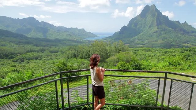 Single women at a look-out rail in Bora Bora looks out at the green volcanic peaks before her with blue sky and white clouds in the background on a sunny tropical day in Tahiti.