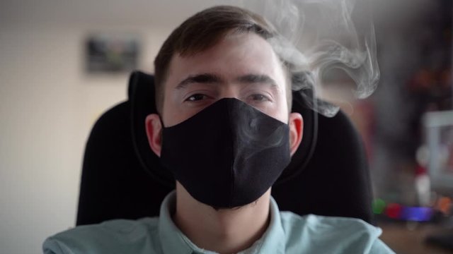 A young man exhales beautifully white marijuana smoke through a black mask.