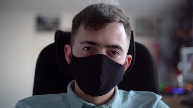 A young guy in a black mask exhales white smoke. The look is directed to the camera.