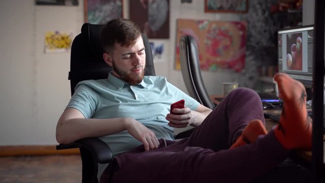 A young bearded guy smokes marijuana while sitting on an armchair. Hemp socks are on the table.