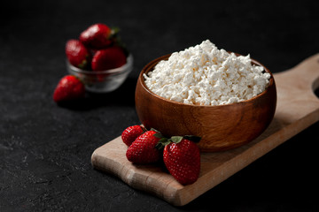 fresh homemade cottage cheese in a wooden plate with strawberries on a cutting board, healthy breakfast on a black table