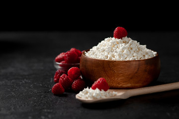 fresh homemade cottage cheese in a wooden bowl with raspberries on a dark background, healthy food on a black table