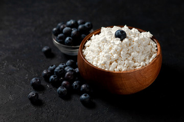 homemade cottage cheese with blueberries in a wooden plate against a dark background, healthy milk breakfast with berries on a black table