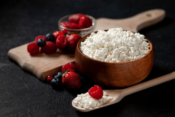 homemade cottage cheese without additives in a wooden bowl with strawberries, raspberries, blueberries, currants against a dark background, healthy milk food with wild berries on a black table