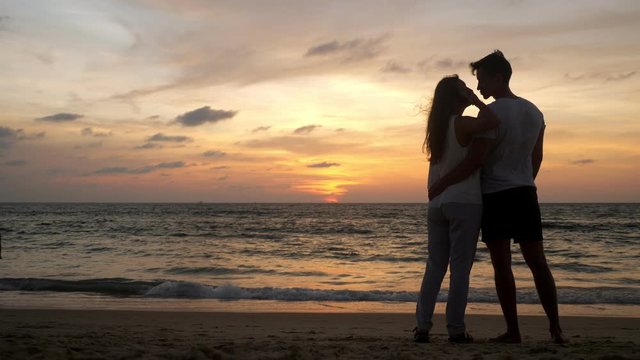 silhouette of young couple spending time on ocean coast joining hands at resort against sunset on horizon backside view