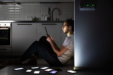 Mobile addiction, nomophobia, insomnia, sleep disorder. Bored man using tablet at late night in the kitchen, chatting on social networks, sitting on the floor around the set of switched-on phones.