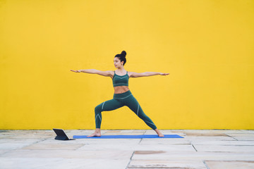 Fototapeta na wymiar Adult fit female practicing yoga in warrior pose using tablet
