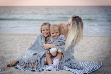 Mom with daughters blondes in white dresses laugh, hug and sit near the blue sea on the beach at sunset.