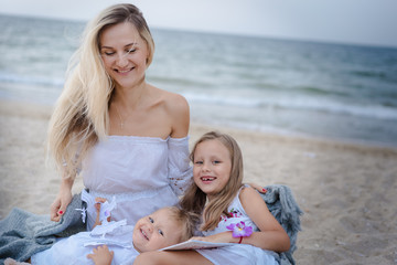 Mom with daughters blondes in white dresses laugh and sit near the blue sea on the beach at sunset. Mom and daughter hold a garland of paper dolls.
