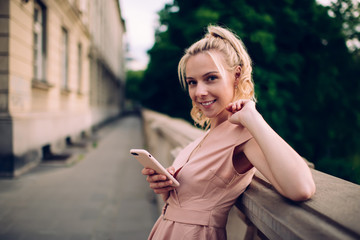 Playful woman in dress texting on smartphone while leaning on parapet of street terrace