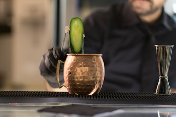 Close up on a copper mug on a bar desk, bartender is pouring a cocktail into it; background is...