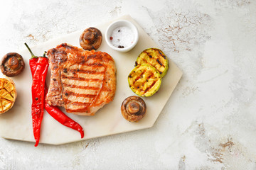 Board with cooked pork steak and vegetables on white background