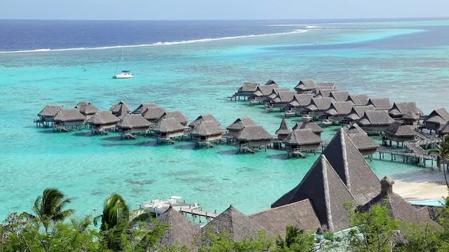Over-water bungalows in Bora Bora seen from the cliffs above showing the clear turquoise water revealing the white sand below as a single lazy catamaran anchors nearby on a sunny tahitian days
