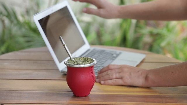 Woman working on laptop and drinking yerba mate tea in home office. Female hands typing on laptop, drinking hot yerba mate tea. Slow Motion.