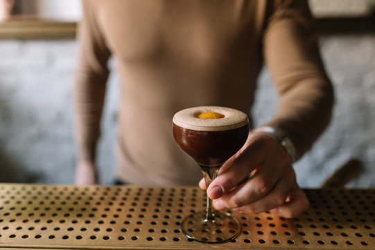 Bartender holding on his hand glass with espresso martini cocktail decorated with slice of orange zest. Smooth image with shallow depth of field.