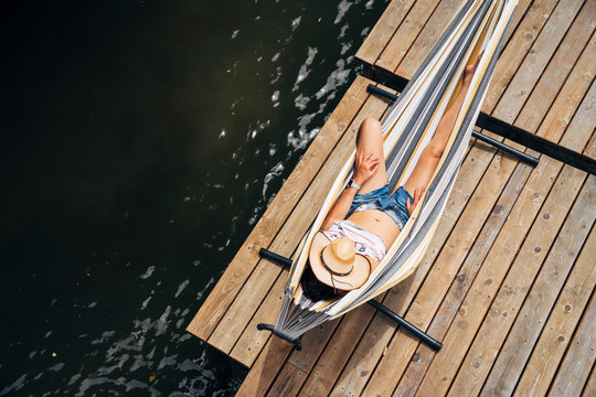 High angle view of woman's face covered with hat relaxing in hammock on dock