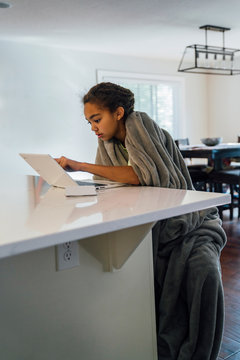 Girl wrapped in blanket studying on touch screen laptop at kitchen counter