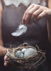 Woman hands holding nest with marble Easter eggs with feathers on dark background. Happy Easter holiday, selective focus, toning