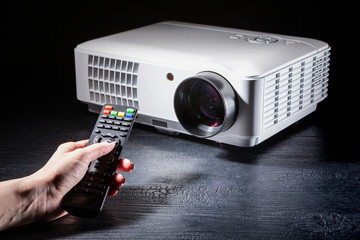 Projector stands on a wooden table. Hand holds the remote control from the projector. Concept -...