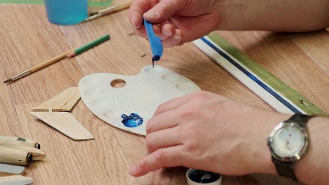 mature man puts blue paint onto palette making jetliner model at table in Sunday school classroom closeup slow motion