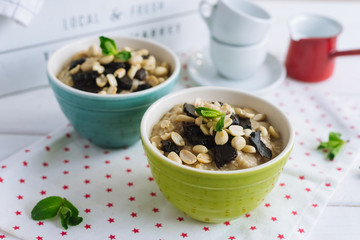 Colored tall bowls with oatmeal with peanuts, prunes, mint leaves. On a white cloth napkin with little red stars. On a white wooden table with cups in the background.