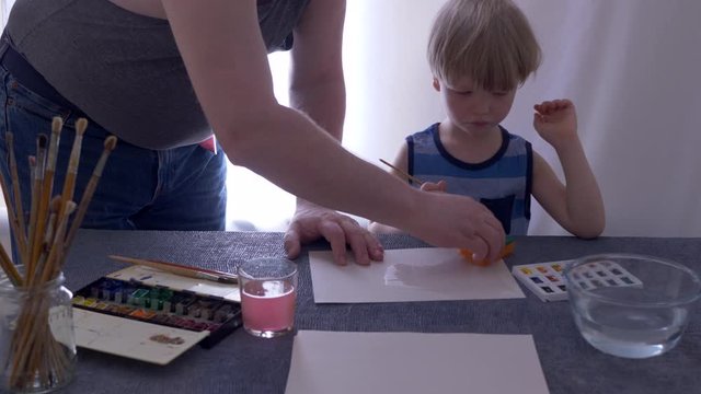 adult father teaches to paint lovely blond child with watercolors. Paints in cuvettes, brushes, water for wetting paper. boy likes to spend time with his father. Wet paper produces beautiful patterns