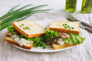 White wheat bread toasted sandwich with pork meat and cheese, salad and sour cream. Chopped chives, bottled olive oil, a knife and a fork. Against the background of a gray fabric napkin.