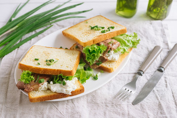 Fototapeta na wymiar White wheat bread toasted sandwich with pork meat and cheese, salad and sour cream. Chopped chives, bottled olive oil, a knife and a fork. Against the background of a gray fabric napkin.