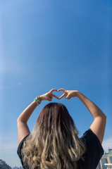 Young asian girl with dyed gray curvy hair and bracelet showing love heart shape figure with hand finger in the background of cityscape and the blue sunny sky