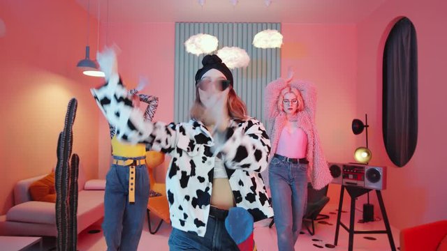 Zoom out shot of three cool female dancers in trendy outfits performing hands movements and dancing femme vogue in vintage studio with creative interior and pink lighting