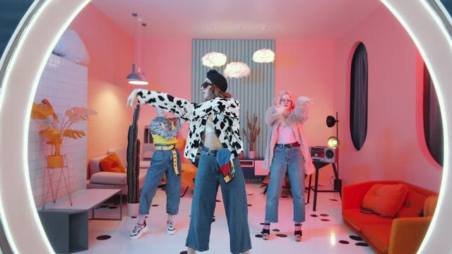 Zoom out shot of three eccentric young ladies in chic outfits performing hands movements and dancing femme vogue for camera in retro styled studio with pink lighting