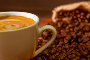 Close-up shot of hot fresh coffee or cappuccino for the morning with milk foam in a white ceramic cup with dark brown coffee beans roasted fragrance in a sack on table background with copy space.