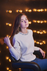 A beautiful young girl is sitting on a chair against the background of a wall with light bulbs