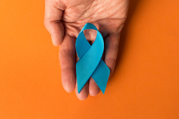 Blue ribbon in human hand on a orange background.  The concept of cancer awareness. Close up. Empty...