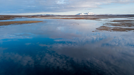calm water reflects blue sky with white clouds against brown meadows and snowy hillpeak on horizon