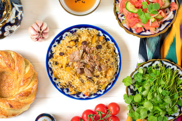 National Uzbek pilaf with meat, achichuk salad of tomato, cucumber, onion in plate with traditional pattern, cilantro, cherry tomatoes, garlicbread tortilla - patir on white wooden table Top view