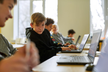 Seeking help. Portrait of caucasian schoolboy looking at the laptop of his neighbor while sitting...