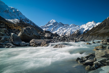 Fototapeta na wymiar Glacier water flowing down a river surrounded by jagged rock formations and snow covered mountains in the background at Hooker Valley glacier at Mount Cook in New Zealand