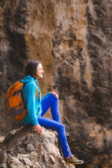 A woman with a backpack sits on a large stone on a background of rocky mountains.