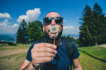 Frontal portrait of a handsome young bearded man blowing a dandelion flower in a high outdoor...