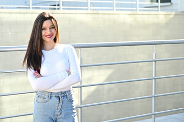 Caucasian portrait of a pretty brunette girl in a white blouse and blue jeans stands near the railing with a smile against the background of a gray wall of a building on a sunny spring day.