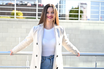 Caucasian portrait of a lovely brunette girl in a light coat and blue jeans with a smile stands near the railing on a sunny spring day.